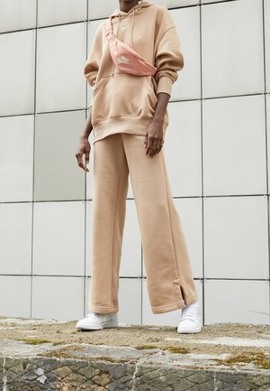 Osoba, která má na sobě béžovou oversized mikinu a odpovídající široké kalhoty, bílé tenisky, stojící u obkládané stěny, s růžovou crossbody taškou Nike.