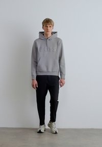 Hall halliga kapuutsiga dressipluus, mille esiküljel on reljeefne "BOSS" logo, esitasku ja soonikkoes mansetid, kombineeritud mustade pükste ja valgete spordijalatsitega.