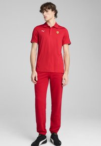 Rød polo-shirt med korte ærmer, udstyret med et Ferrari-logo og hvide accenter. Parret med matchende røde bukser og sorte sneakers.