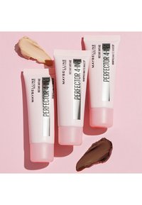 Tre tubetti di fondotinta Maybelline Perfecter 4-in-1 Whipped Matte con confezione rosa e bianca, accompagnati da campioni di trucco beige e marroni su uno sfondo rosa.