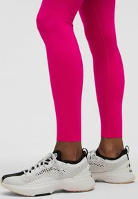 Fehér és fekete sportcipők hálós részletekkel és vastag talppal, összeillő, élénk rózsaszín leggings-szel párosítva.