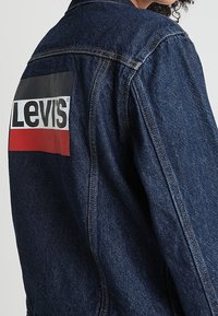 Persona che indossa una giacca di jeans blu scuro con un grande patch rettangolare del logo Levi's in nero, bianco e rosso sulla schiena.