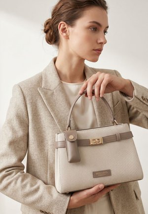 Femme en blazer beige tenant un sac à main beige et taupe texturé avec un fermoir doré et une petite pochette attachée.