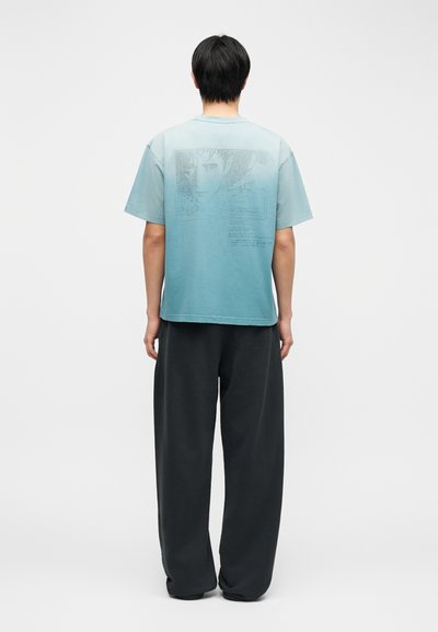 Άνδρας που στέκεται γυρισμένος με την πλάτη, φοράει μπλουζάκι με διαβαθμισμένο ανοιχτό γαλάζιο χρώμα και διακριτικό γραφικό στο πίσω μέρος, μαζί με άνετο μαύρο παντελόνι, σε απλό φόντο.