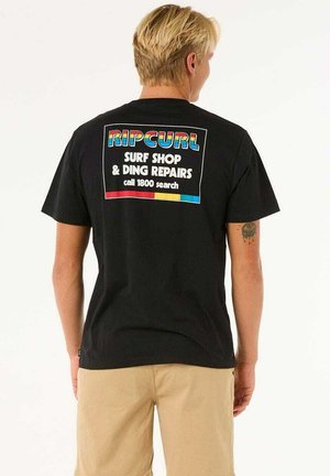 Camiseta de algodón negra con un gráfico colorido en la parte posterior que dice "RIPCURL SURF SHOP & DING REPAIRS" y detalles de contacto centrados dentro de un recuadro.