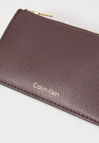 Calvin Klein novčanik od teksturirane burgundske kože s zlatnim logotipom, elegantnog pravokutnog oblika i zatvaranjem na patentni zupčanik.