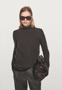 Ciemnobrązowy prążkowany sweter z golfem z guzikami na ramionach, noszony na czarnej satynowej warstwie, w połączeniu z szarymi plisowanymi spodniami i brązową torbą.