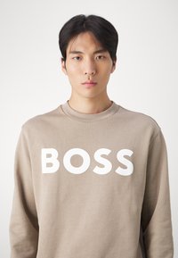 Sudadera beige con cuello redondo y mangas largas. En la parte frontal se muestra la palabra "BOSS" en grandes letras blancas. Textura suave.