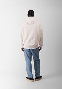 Lys pink hoodie med løs pasform og stor baglomme, parret med lyseblå jeans og sorte sko, set fra bagen.