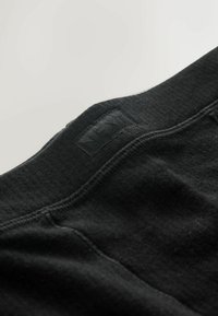 Tela negra con costillas y una cinturilla cosida que presenta una etiqueta negra elevada que dice "NEXT". Textura suave con detalles de costura sutiles.