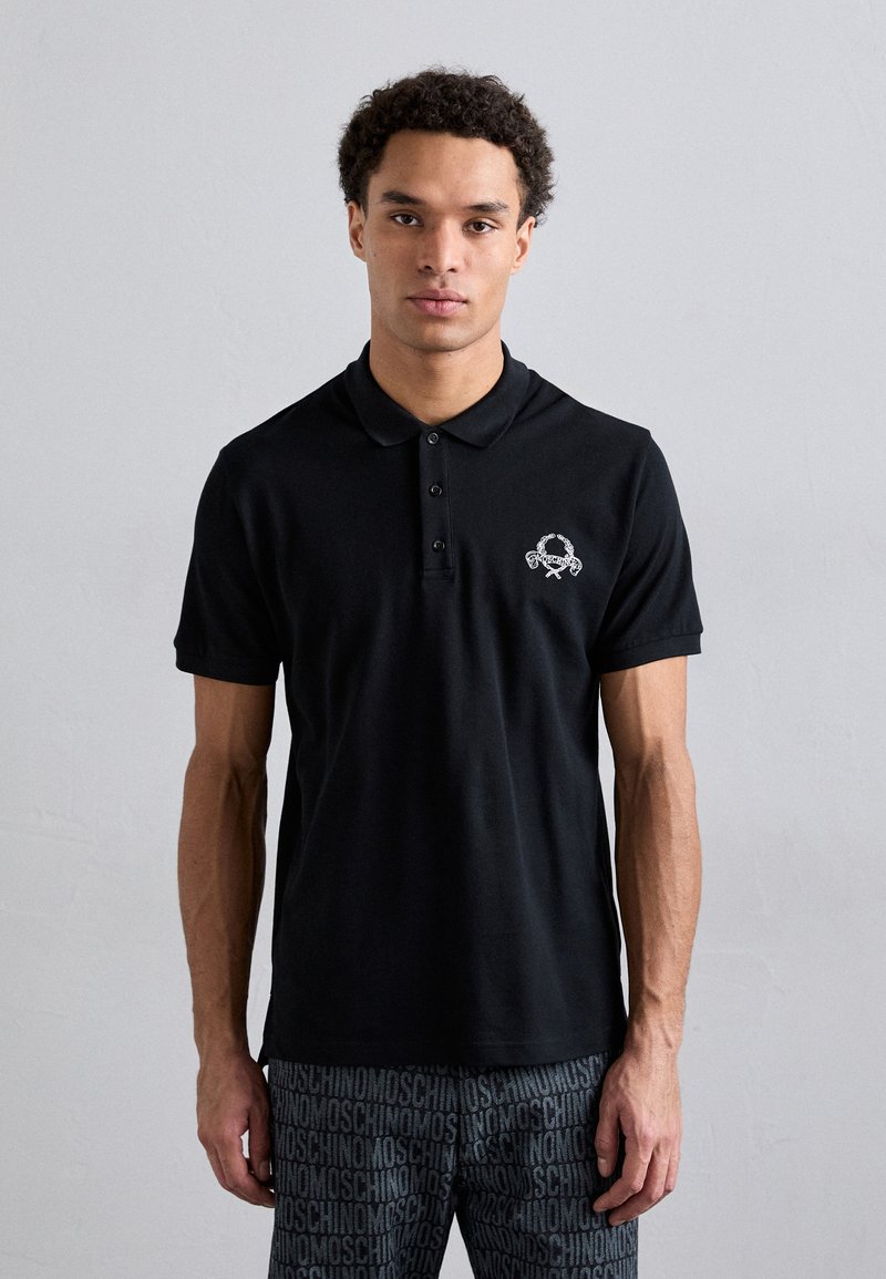 Svart polo skjorta i bomull, med krage och placket med tre knappar. Broderad design på vänster bröst. Korta ärmar.