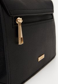 Bolso negro texturizado con un tirador de cremallera dorado y detalles en dorado. Presenta una forma rectangular suave y un bolsillo delantero con una placa de logo.