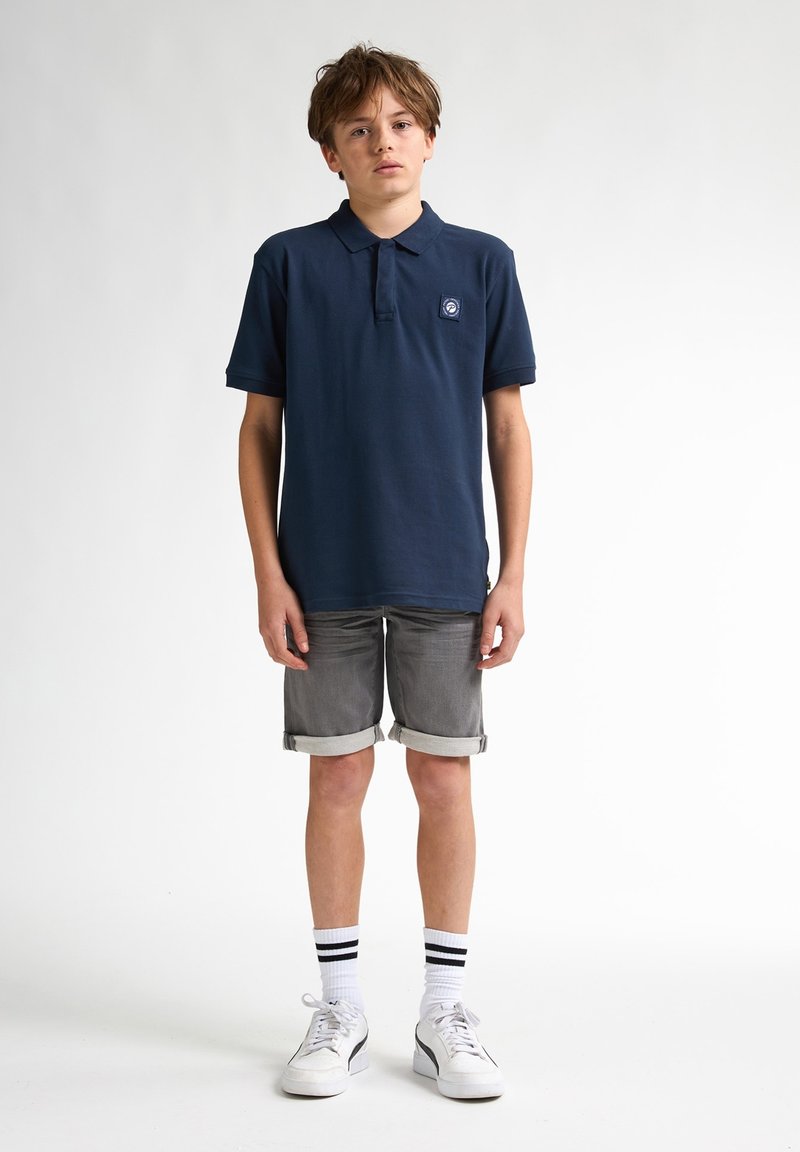 Marinblå polo, gråta rullade shorts, vita sneakers med svarta accenter och knälånga vita strumpor med svarta ränder. Enkel, avslappnad design.