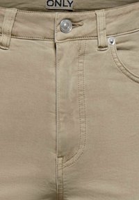 Pantalon en denim beige avec bouton à l'avant, fermeture éclair, poche avant, et passants pour ceinture, arborant une petite étiquette de marque "ONLY" à la ceinture.