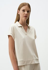 Beige Poloshirt mit V-Ausschnitt, kurzen Ärmeln und lockerer Passform. Hergestellt aus weichem Material mit glatter Textur und dezentem Glanz.