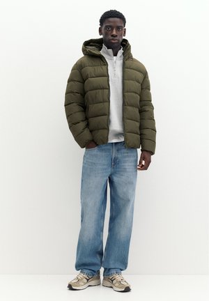 Jeune homme portant une doudoune vert olive, un pull gris clair à zip quart, un jean bleu ample et des baskets beiges, debout devant un fond blanc.