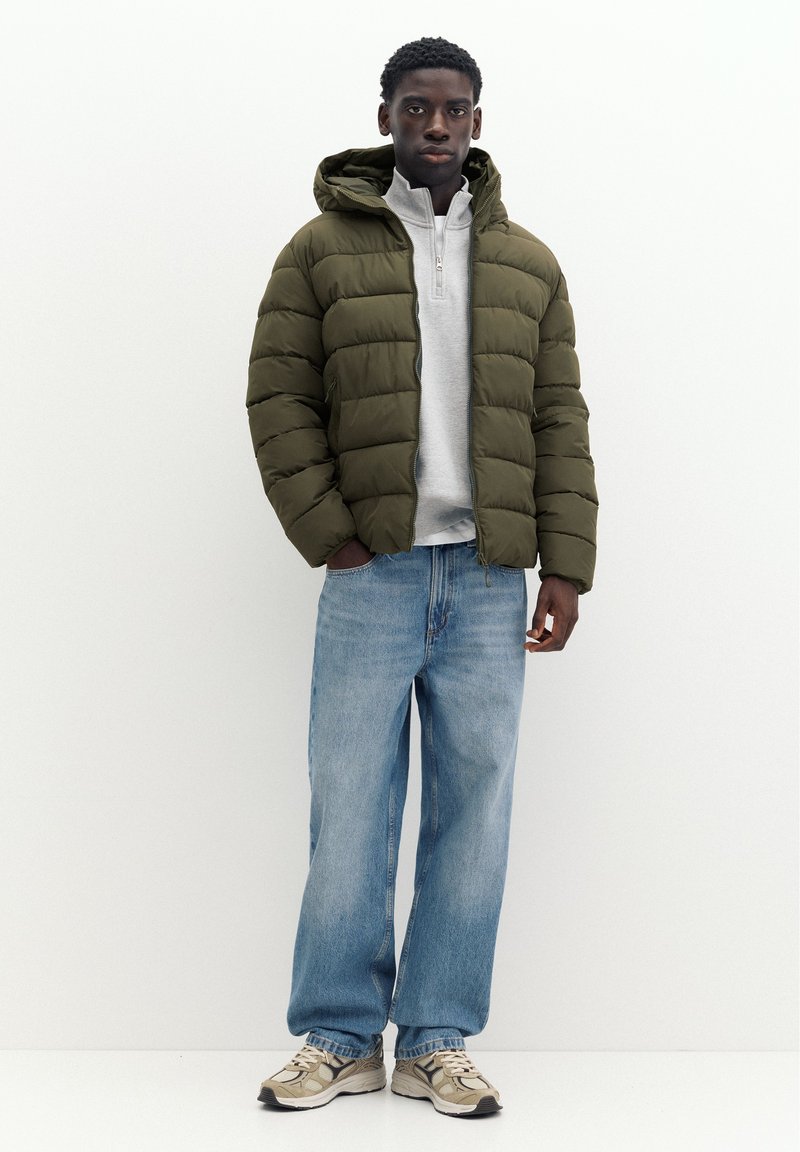 Jeune homme portant une doudoune vert olive, un pull gris clair à zip quart, un jean bleu ample et des baskets beiges, debout devant un fond blanc.