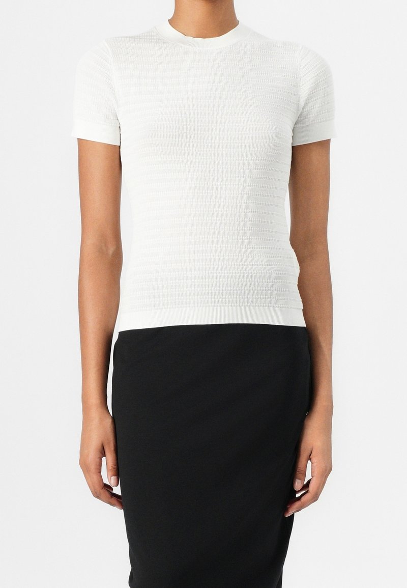 Femme portant un haut en maille blanc texturé à manches courtes associé à une jupe noire taille haute, montrée du cou aux hanches.