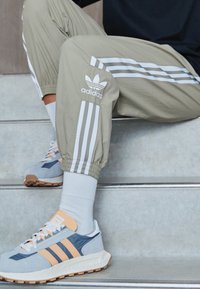 Person som sitter på trappor iförd beige Adidas träningsbyxor med vita ränder och blå och orange Adidas sneakers med vita strumpor.