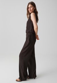 Woman with long brown hair wearing a sleeveless black jumpsuit and sandals, standing sideways with hands clasped behind back.