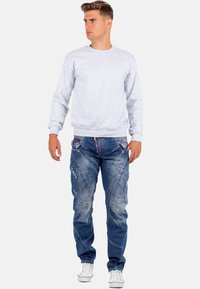 Felpa grigia a girocollo abbinata a jeans in denim blu strappati con cuciture uniche e tasche con cerniera. Sneakers bianche completano il look.