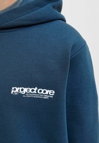 Felpa con cappuccio di colore blu scuro, realizzata in tessuto morbido, con un logo bianco "project core" e testo, caratterizzata da un design spazioso e con cappuccio.