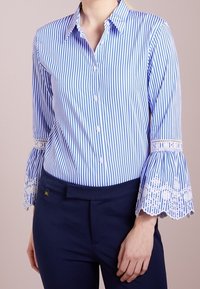 Randig blå och vit knapp-up skjorta med utsvängda ärmar som har spetsdetaljer, i kombination med mörka byxor. Skräddarsydd passform i bomullsmaterial.