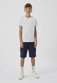 Maglietta bianca in cotone, pantaloni cargo blu navy con tasche laterali, sneakers bianche e calzini bianchi con logo, in piedi contro uno sfondo neutro.