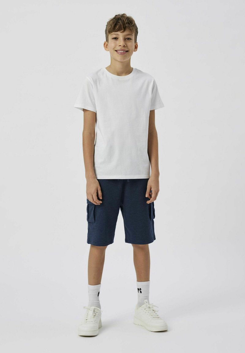 Maglietta bianca in cotone, pantaloni cargo blu navy con tasche laterali, sneakers bianche e calzini bianchi con logo, in piedi contro uno sfondo neutro.