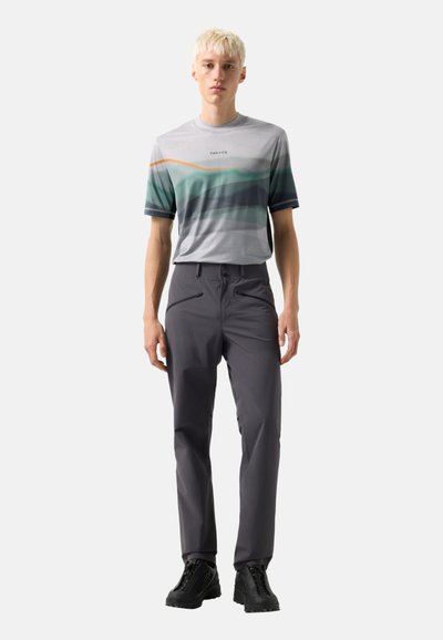 Junge Person mit kurzen blonden Haaren, die ein graues T-Shirt mit Grafik, dunkelgraue Hosen mit Reißverschlusstaschen und schwarze Wanderschuhe trägt, stehend.