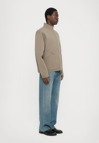 Chaqueta beige con cremallera y cuello alto, con dos bolsillos delanteros con cremallera, llevada sobre unos vaqueros anchos de color azul claro y zapatos negros. Textura de tela suave.
