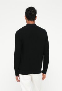Suéter negro de punto con un acabado texturizado, que presenta un cuello y mangas largas, combinado con pantalones de color claro.