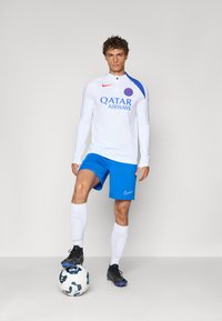 White long-sleeve soccer jersey with blue accents, "QATAR AIRWAYS" logo, blue shorts, white socks, standing on a soccer ball, black cleats.