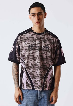 Joven con cabello corto y oscuro que lleva una camisa con estampado de camuflaje en morado y negro con detalles gráficos y jeans azules, de pie frente a un fondo liso.