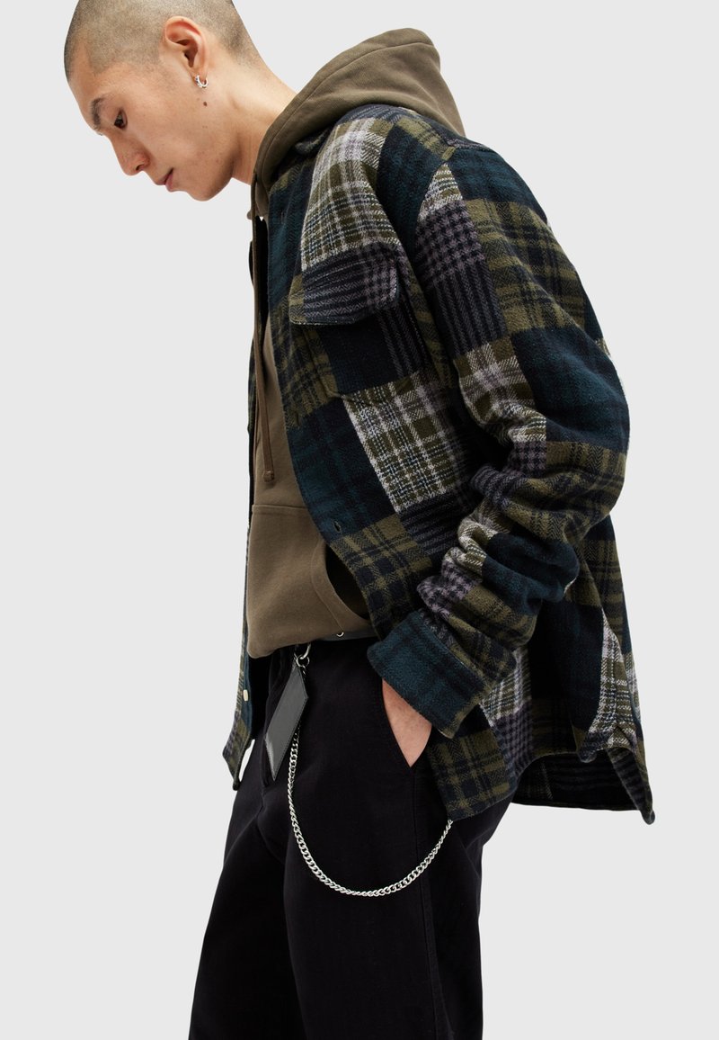 Flanellen overhemd met een gemengd ruitpatroon in donker groen, marineblauw en grijs, gecombineerd met een bruine hoodie. Zwarte broek met een zilverkleurige kettingdetail.