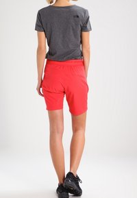 Graues T-Shirt mit kurzen Ärmeln und einem Logo auf dem Rücken, kombiniert mit leuchtend roten Sportshorts. Schwarze Turnschuhe vervollständigen das Outfit.