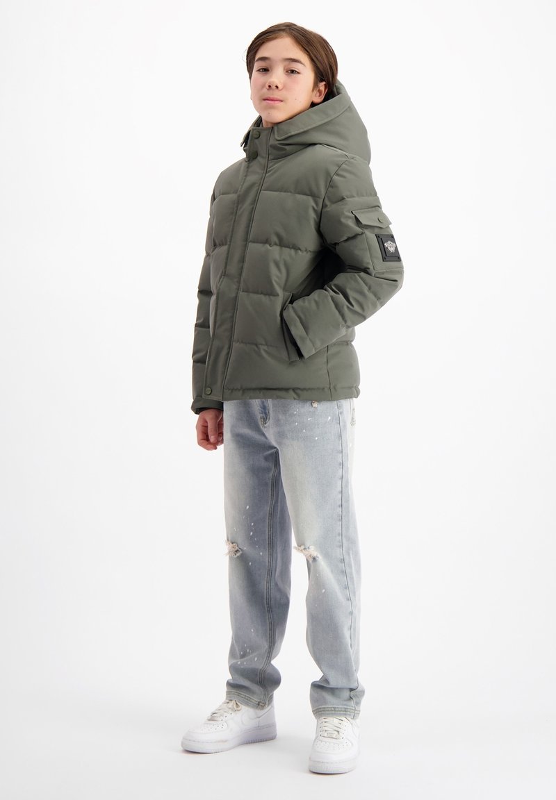 Groene pufferjack met capuchon, voorzien van een geborduurd ontwerp en zijzakken. Gecombineerd met lichtblauwe versleten jeans en witte sneakers.