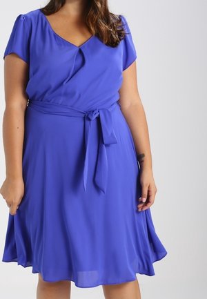 Woman wearing a knee-length royal blue dress with short sleeves, a V-neckline, and a tied waist belt.