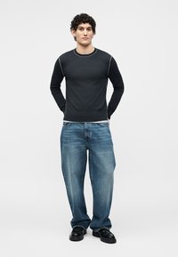 Jeune homme debout, les mains derrière le dos, portant un t-shirt noir à manches longues, un jean bleu ample et des chaussures noires sur un fond blanc uni.