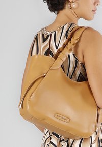 Läderaxelväska i brunt med flätad handtag och logodetalj. Kurvad silhuett med slät yta, kombinerad med en mönstrad klänning.