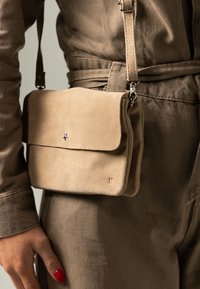 Sac en bandoulière en suède beige avec fermoir en métal argenté porté sur une tenue en denim taupe ceinturée, main avec vernis à ongles rouge reposant à proximité.