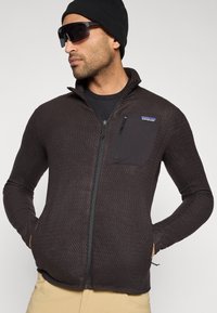 Chaqueta polar negra con cremallera y patrón texturizado, que incluye un bolsillo en el pecho y bolsillos laterales. Se lleva sobre una camisa negra, con un gorro y gafas de sol.