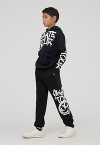 Czarna bluza z kapturem z białym tekstem w stylu graffiti, zestawiona z czarnymi spodniami dresowymi z dużym białym wzorem. Całość dopełniają białe sneakersy.