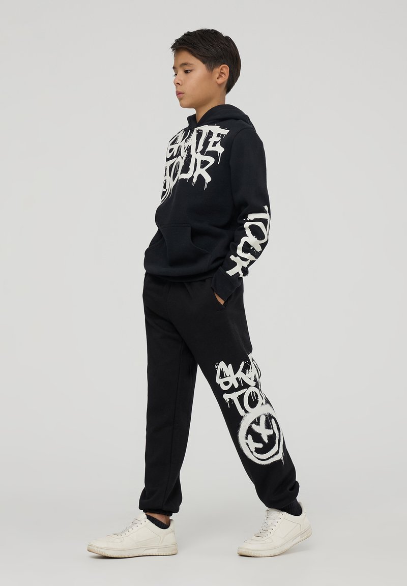 Czarna bluza z kapturem z białym tekstem w stylu graffiti, zestawiona z czarnymi spodniami dresowymi z dużym białym wzorem. Całość dopełniają białe sneakersy.