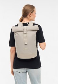 Person with light brown hair in ponytail wearing black shirt and light denim jeans carries beige roll-top backpack with strap buckle.