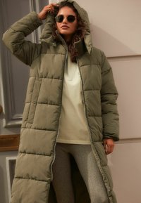 Manteau rembourré long de couleur olive avec une capuche, présentant un motif matelassé, une fermeture éclair frontale et des fentes latérales pour plus de liberté de mouvement.
