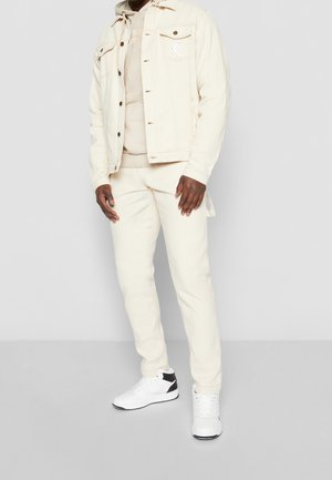 Beige džísiová bunda se záplatou, béžová mikina a odpovídající kalhoty. Bílé tenisky s černými akcenty. Hladká textura, pasovaný styl.