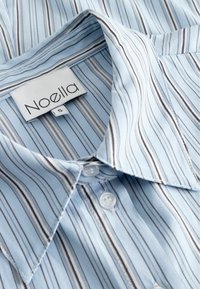 Világoskék ing függőleges fehér és tengerész kék csíkokkal. Klasszikus gallérral, gombos elülső részel és "Noella" felirattal ellátott kis méretű címkével rendelkezik.