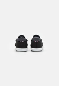 Black canvas sneakers with a white sole, featuring a padded collar and a small striped accent on the back. Simple and low-profile design.