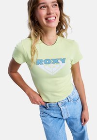 Jasnożółta dopasowana koszulka z niebieskim logo i grafik�ą "ROXY", zestawiona z jasnoniebieskimi dżinsami z wysokim stanem.